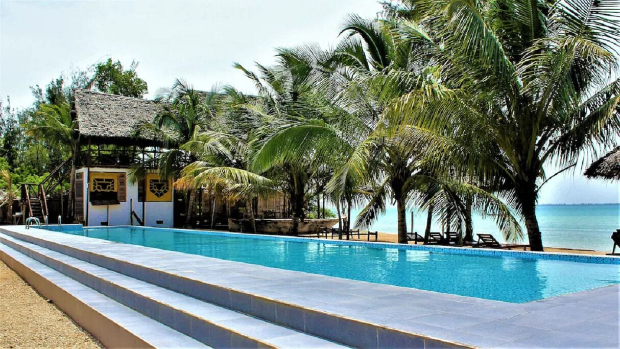 A pool area with a thatched-roof structure, palm trees, and a view of the sea.