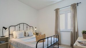 A bedroom with a bed, bedside lamps, a nightstand, a window with curtains, and a small table with a plant. - Dona Ana by Interhome (Lagos)