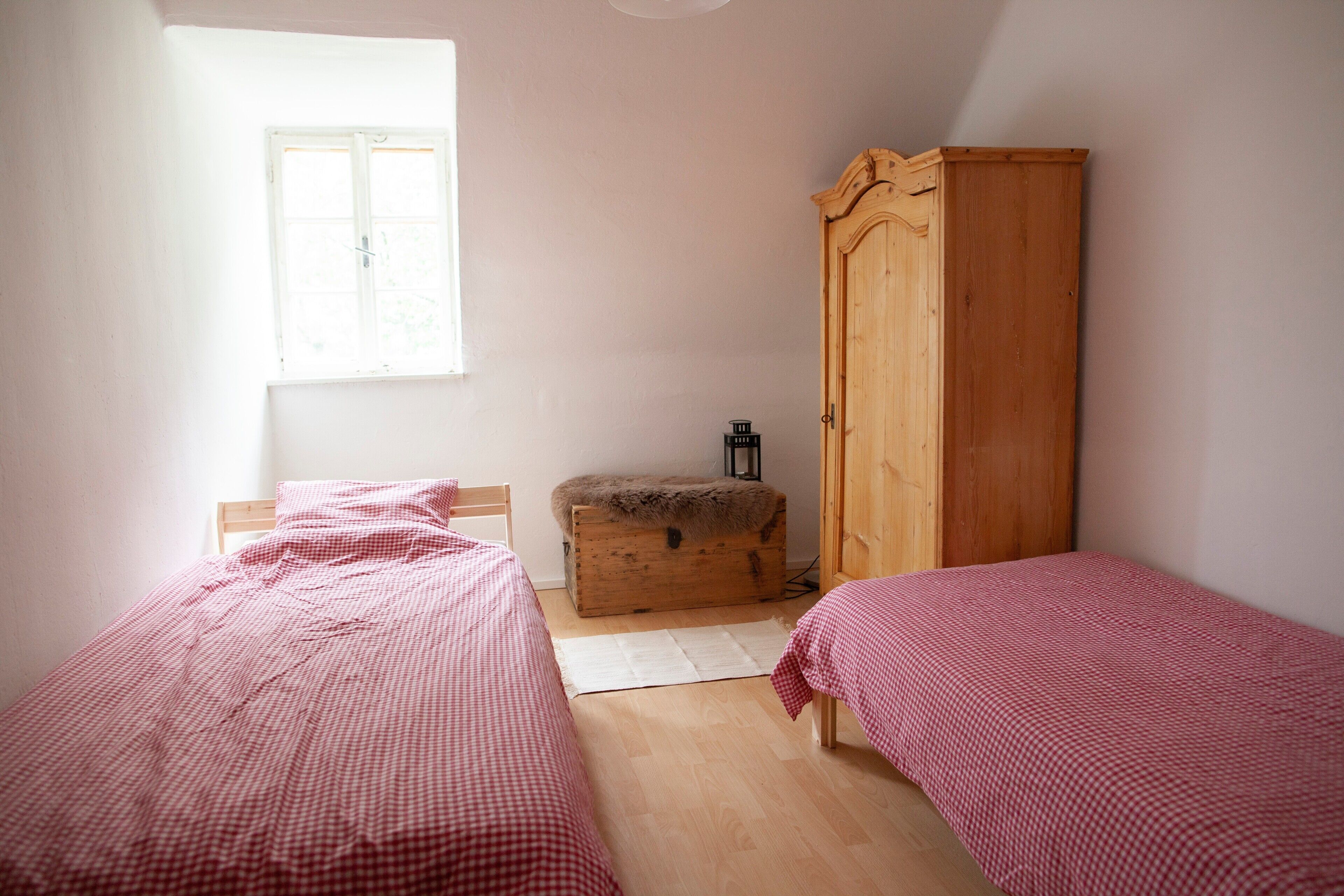 A bedroom with two single beds, a wooden wardrobe, a small chest of drawers, and a window providing natural light.