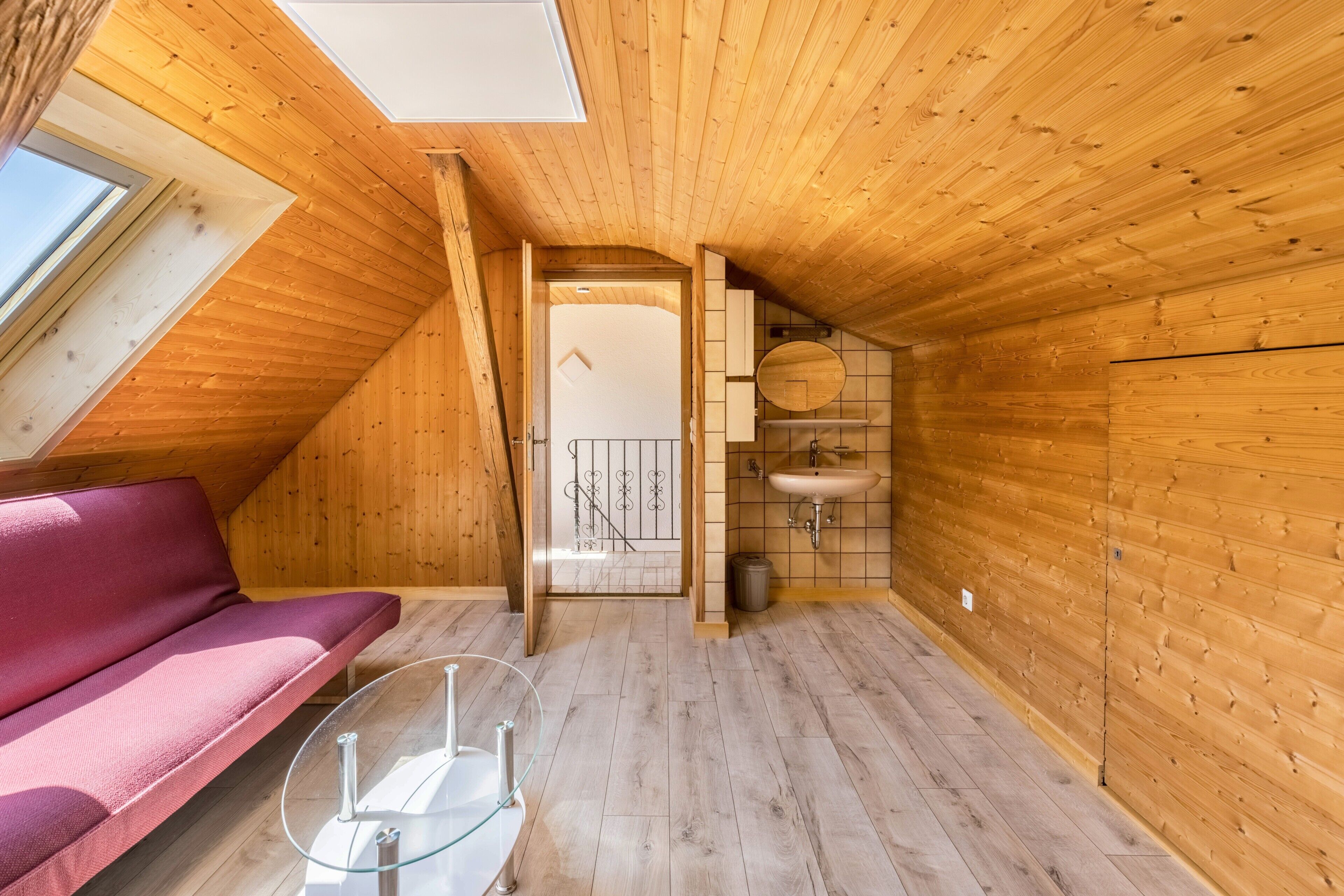 A wooden interior with a sloped ceiling, a red sofa, a glass table, and a bathroom with a sink and toilet.
