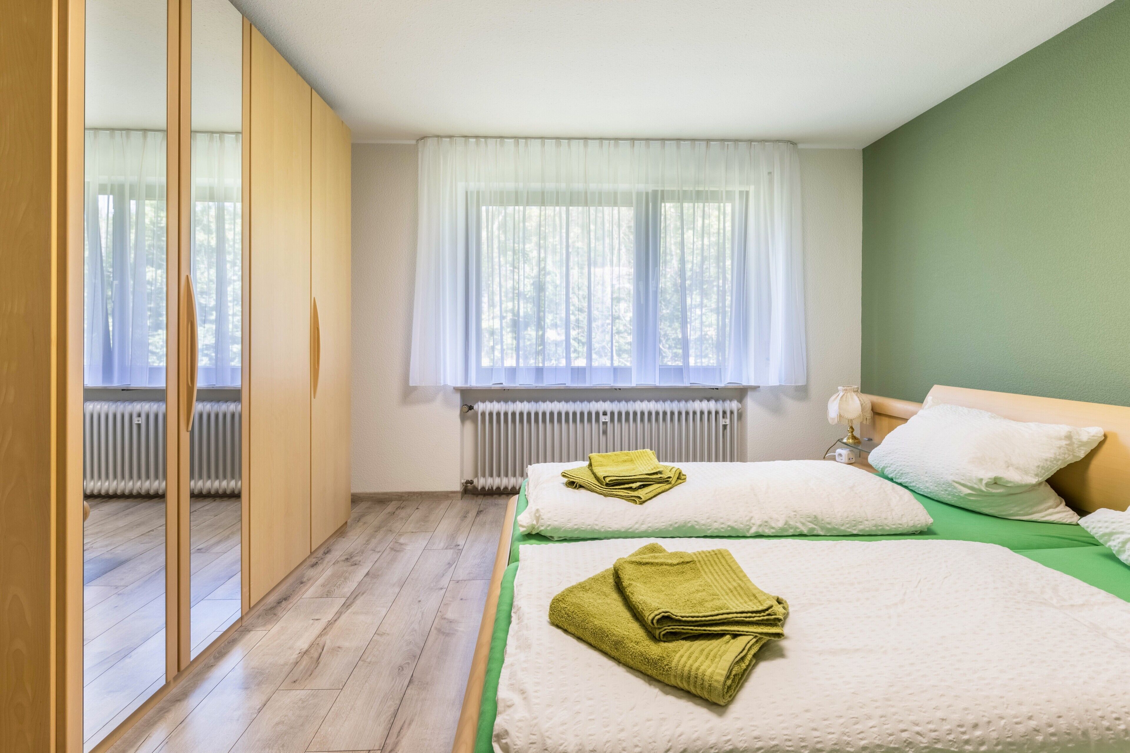 Habitación con dos camas, un radiador, un ventanal con cortinas traslúcidas y suelo de madera.