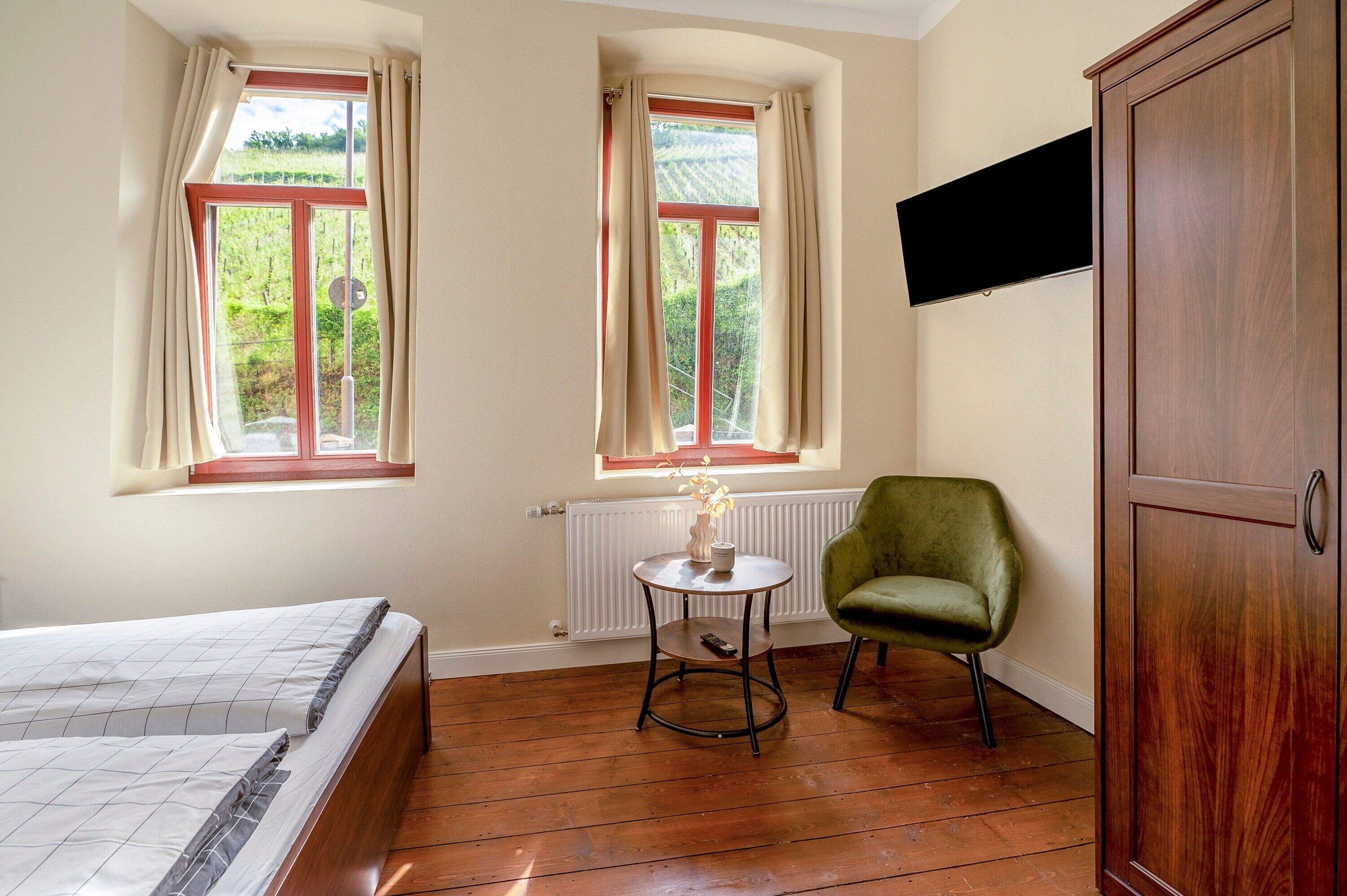A room with a bed, two windows with curtains, a radiator, a small round table with a vase, a green chair, and a wooden wardrobe.