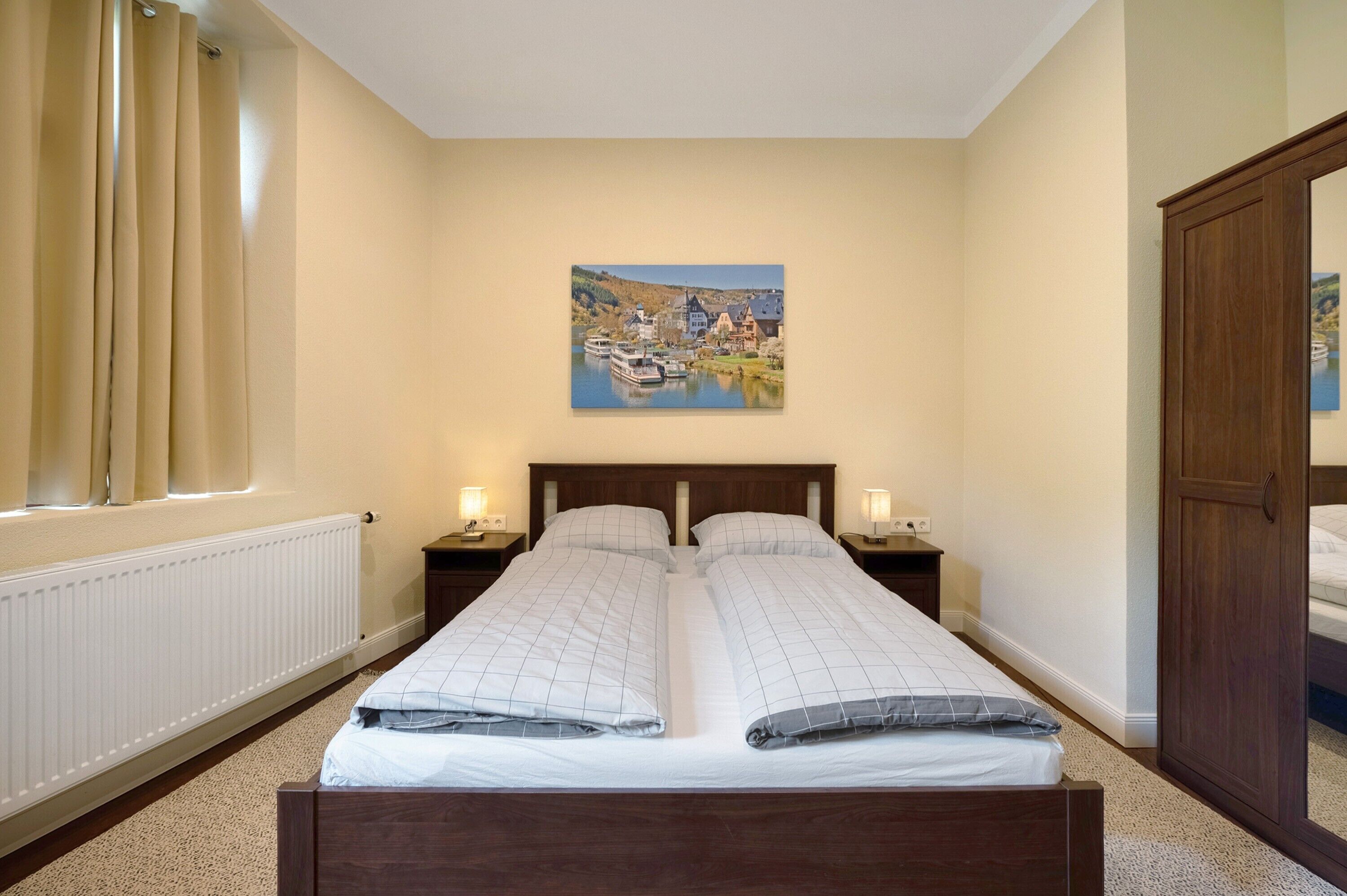 Ein Doppelbett mit Kopfteil, zwei Nachttische, ein Gemälde mit Seeblick, ein Holzschrank und ein Fenster mit Vorhängen.
