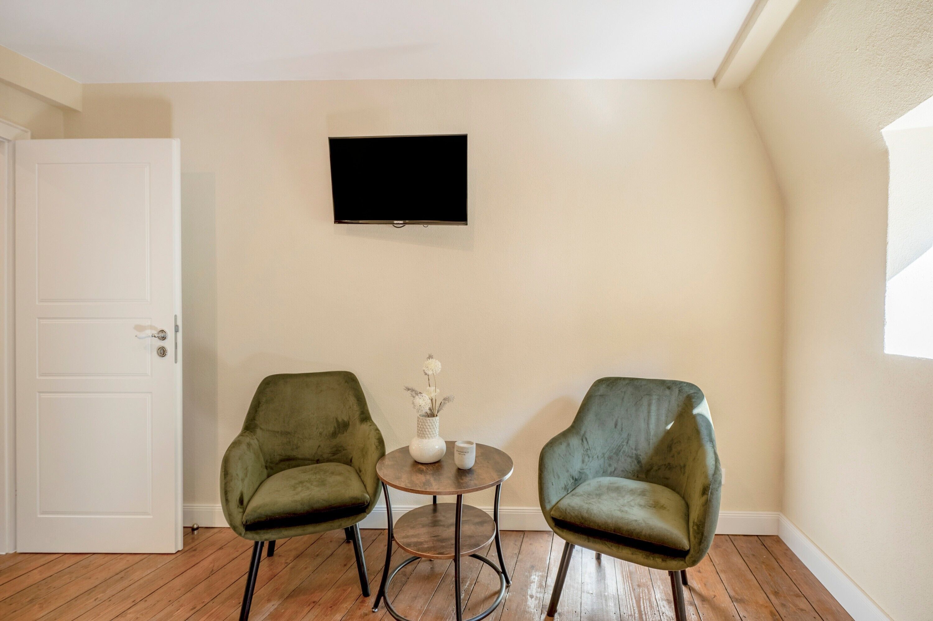 A room with two green armchairs, a small round table with a vase and candle, a flat-screen TV on the wall, and a white door.