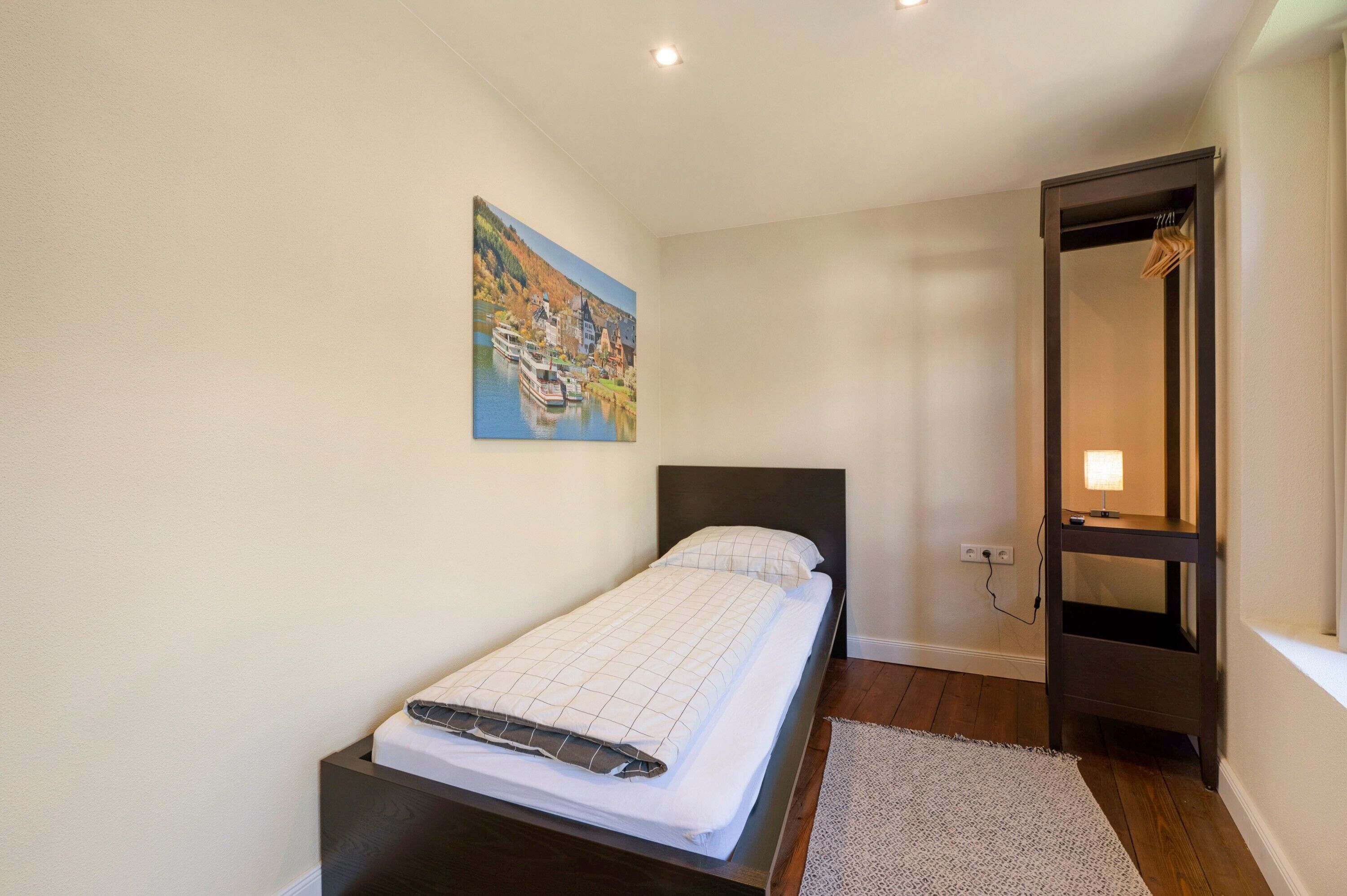 A bedroom with a bed, a painting, a wooden floor, a door, and a small table with a lamp.