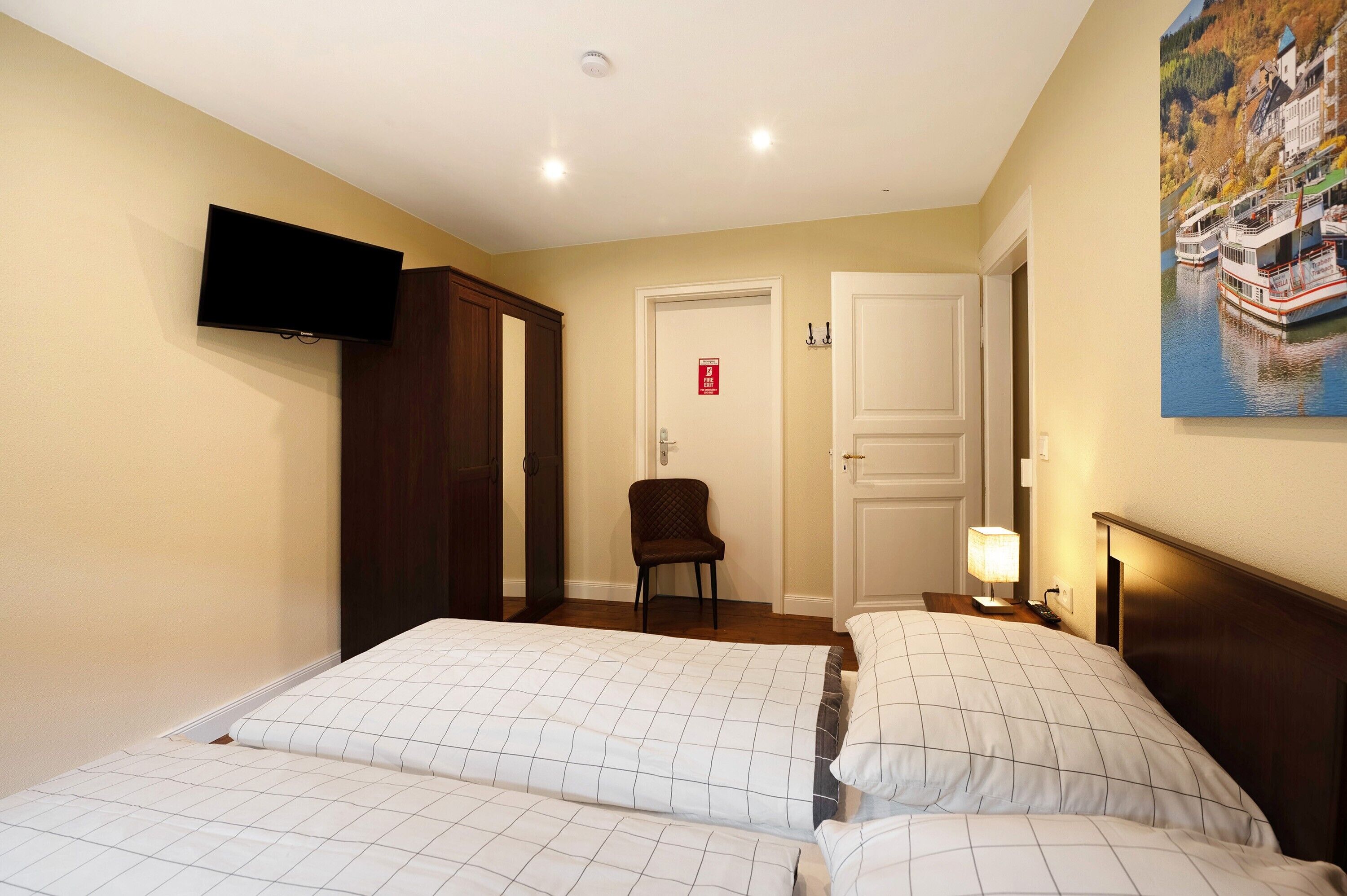 A hotel room with two beds, a TV mounted on the wall, a chair, a door with a fire alarm, and a painting of boats on the wall.