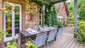 Una terraza de madera con una mesa preparada para comer, rodeada de sillas, y un área cubierta con faroles colgantes.