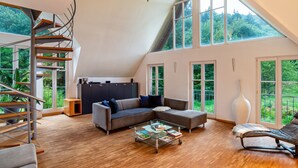 Un loft amplio con una escalera de madera, un sofá seccional, una mesa de centro de vidrio y ventanas grandes que ofrecen vistas a zonas verdes.