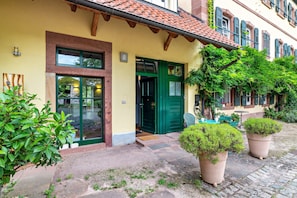 A building with a yellow facade, green doors, and a tiled roof. There are potted plants in front and a cobblestone path.