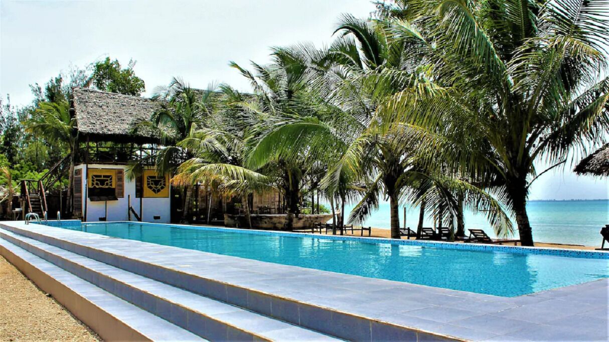 A pool area with a thatched-roof structure, palm trees, and a view of the sea.