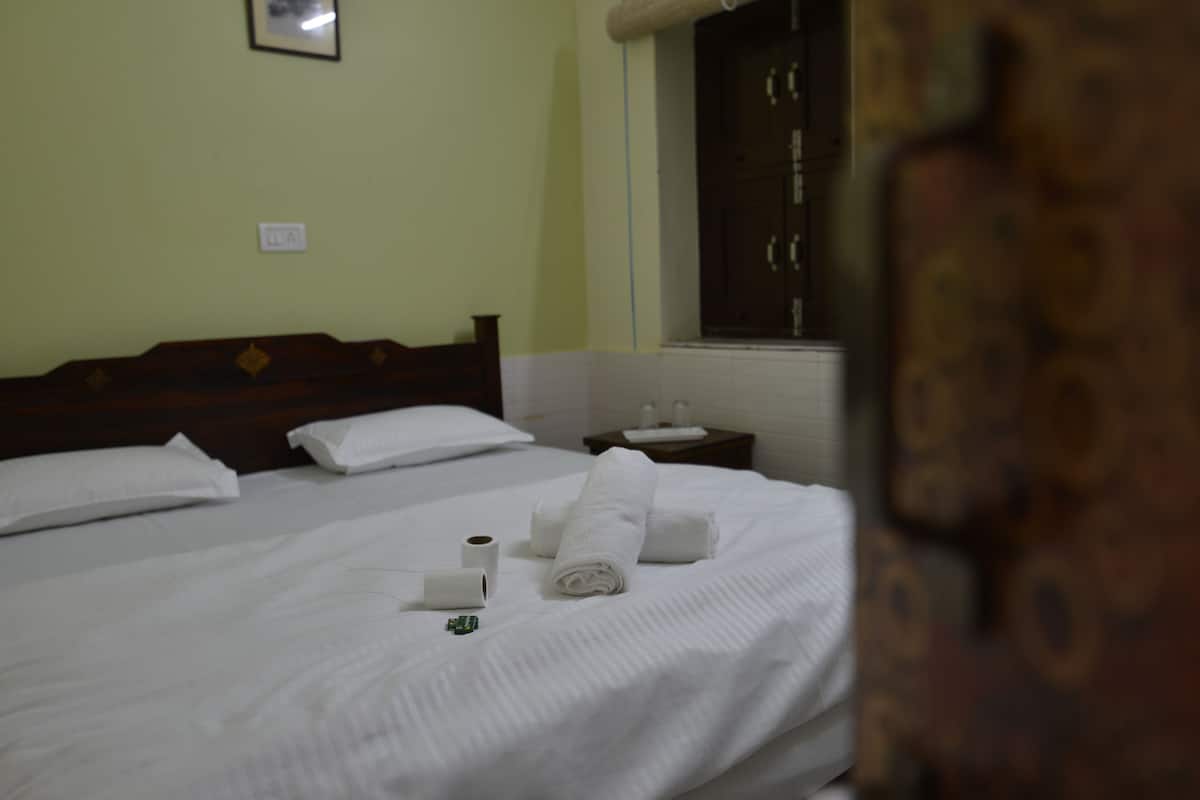Una habitación de hotel con una cama, almohadas, una mesita con una taza y una caja de pañuelos, y una ventana con cortinas.