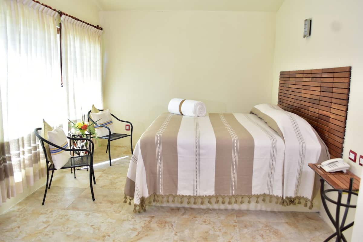 Une chambre d’hôtel avec un lit, une chaise, une petite table avec un téléphone et une fenêtre avec des rideaux.
