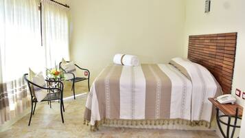 Habitación de hotel con cama, una silla, una mesita con un teléfono y ventana con cortinas.