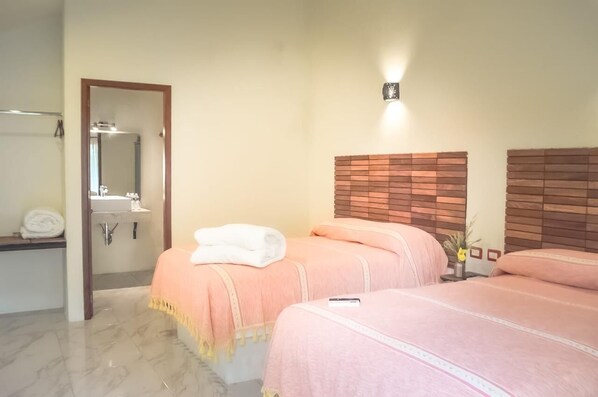 Habitación de hotel con dos camas, un baño con espejo y lavamanos, y un accesorio de iluminación fijado a la pared.