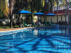 Una piscina con agua cristalina azul, rodeada de palmeras y sillas de descanso bajo sombrillas azules.