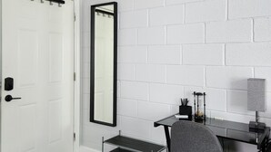Ett modernt rum med en vit tegelvägg, en vit dörr, en stor spegel, ett skrivbord med en stol och en liten hylla.