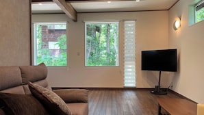 A living room with a brown sofa, a flat-screen TV, and a wooden bench. There are two windows with blinds, one showing a view of trees and the other a brick wall. - BETSUINN MINI (Hakuba)