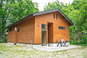 A modern wooden cabin with a large door and windows, a small patio with a table and chairs, and a surrounding forest. - BETSUINN MINI (Hakuba)