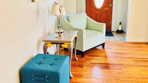 A room with a wooden floor, a blue ottoman, a white sofa, a small table with a lamp, and a door with a circular window.