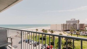 Un balcón con vistas a la playa y al océano, un estacionamiento con coches, y un edificio al fondo.
