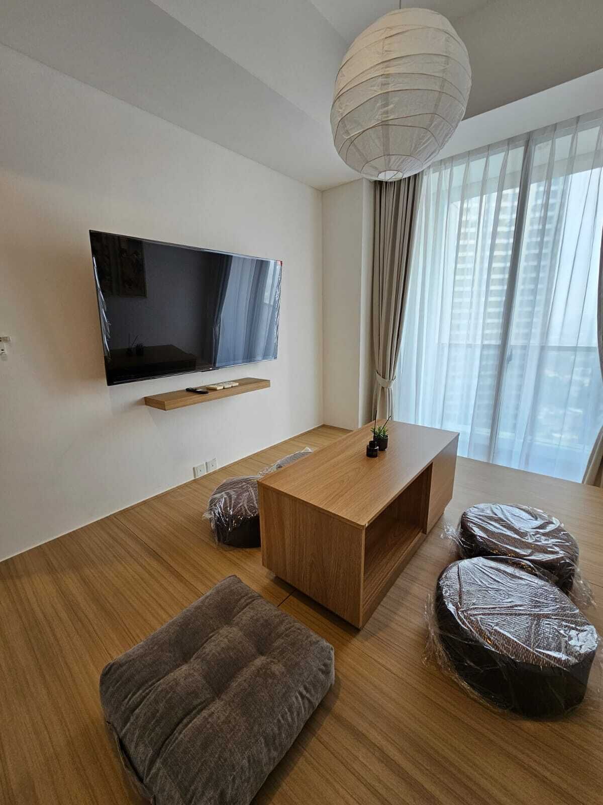 A modern living room with a flat-screen TV mounted on the wall, a wooden coffee table, a brown ottoman, and a large window with sheer curtains.