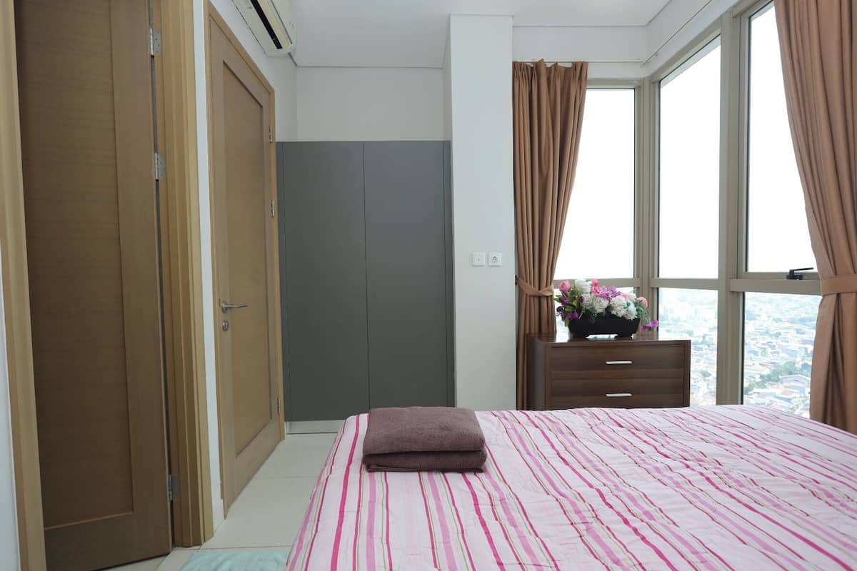 A bedroom with a bed, a dresser, a window with curtains, and a view of the city.