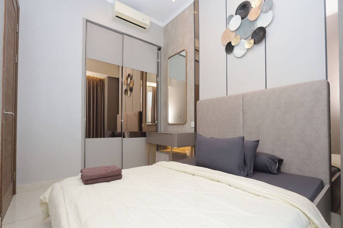 A modern bedroom with a large bed, a wardrobe, a mirror, and a decorative wall piece.