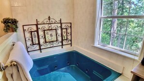 A bathroom with a stone wall, a blue jacuzzi tub, a window with a view of trees, and a decorative metal frame with pictures. - Tuscany Dream Rock Cottage/#8 (Eureka Springs)