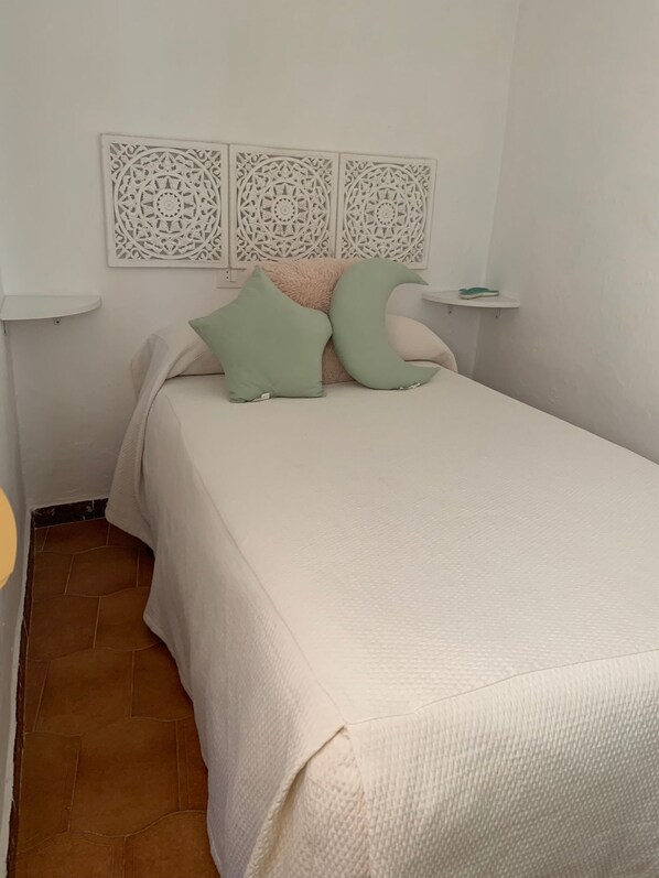 A neatly made bed with a beige bedspread and two green pillows, a white wall with decorative panels, a small white shelf with a green slipper, and a brown tiled floor. - Casa Ana in Vejerde la Frontera (Vejer de la Frontera)
