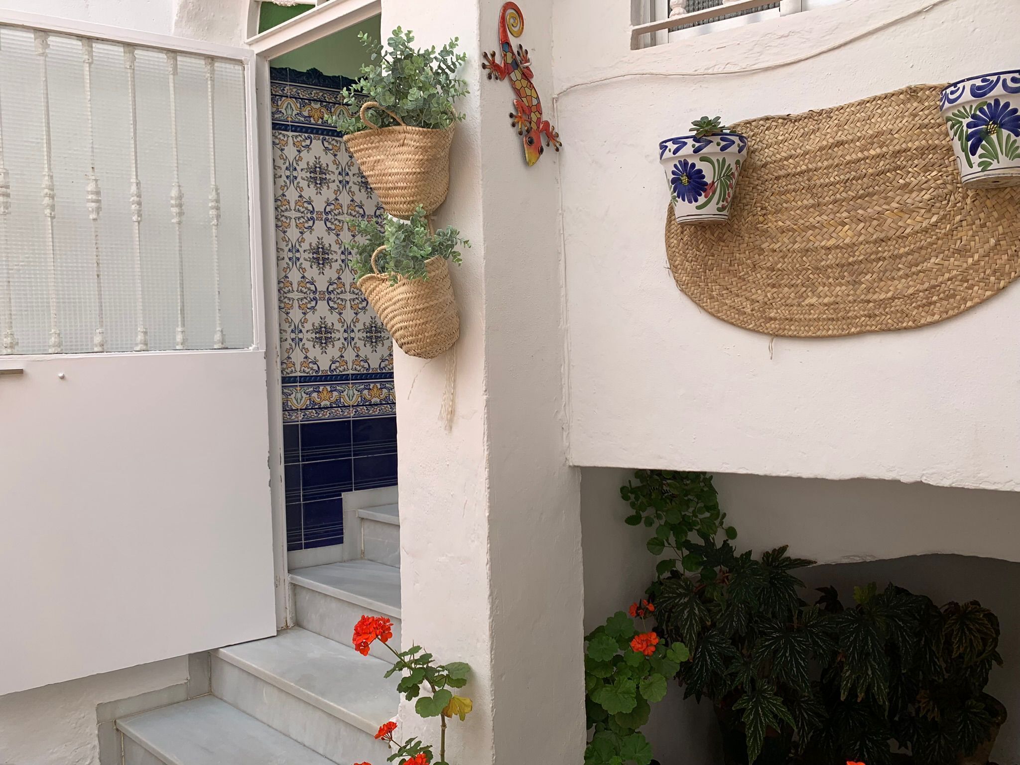 Un escalier aux marches blanches menant à une porte, un mur recouvert de carreaux bleus et blancs formant un motif, et un panier tressé suspendu au mur.