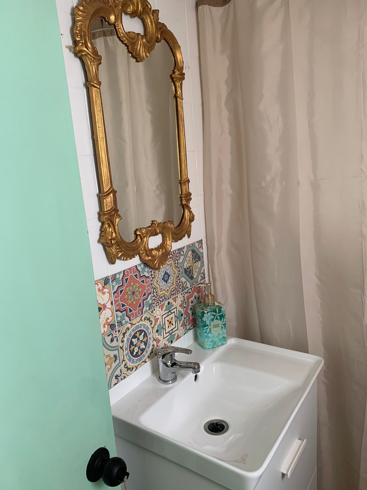 Un bagno con un lavandino bianco, uno specchio dorato decorato e uno schienale colorato in piastrelle.