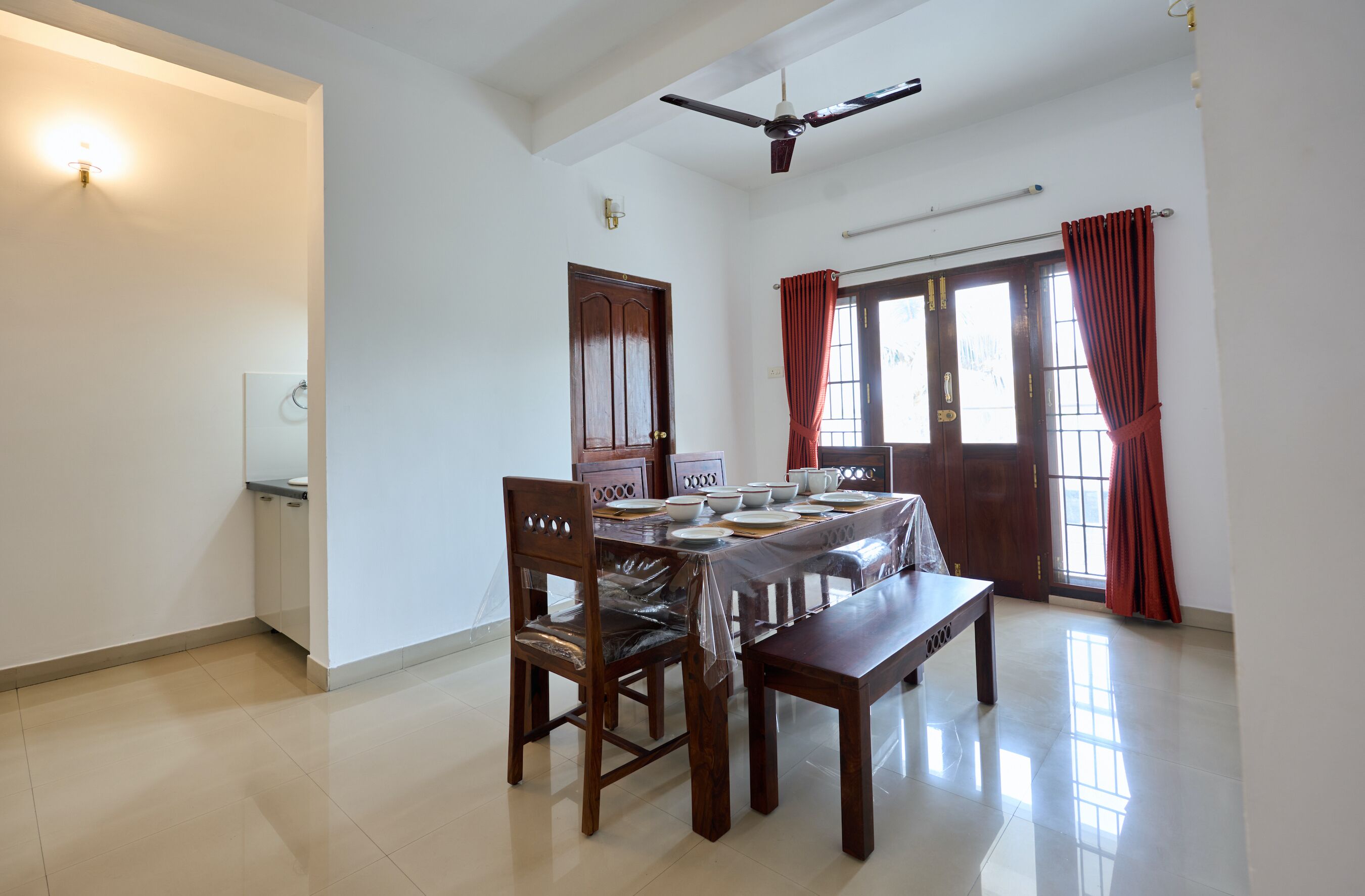A dining area with a table set for four, wooden chairs, a ceiling fan, and a window with curtains.