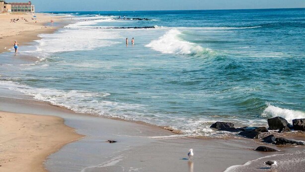 A beach with waves, a few people, and a building in the background. - Best Value, Quality Stay! Close to Tinton Falls Recreation Complex, Pet-Friendly (Tinton Falls)