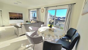 A modern living room with a sofa, chairs, a dining table, and a view of the city through large windows. - Elegant & Convenient New House at Leppington - 2 min drive from train station! (Leppington)