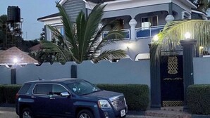 Une maison d’habitation avec une entrée fermée par un portail, un VUS garé et un réservoir d’eau en arrière-plan.
