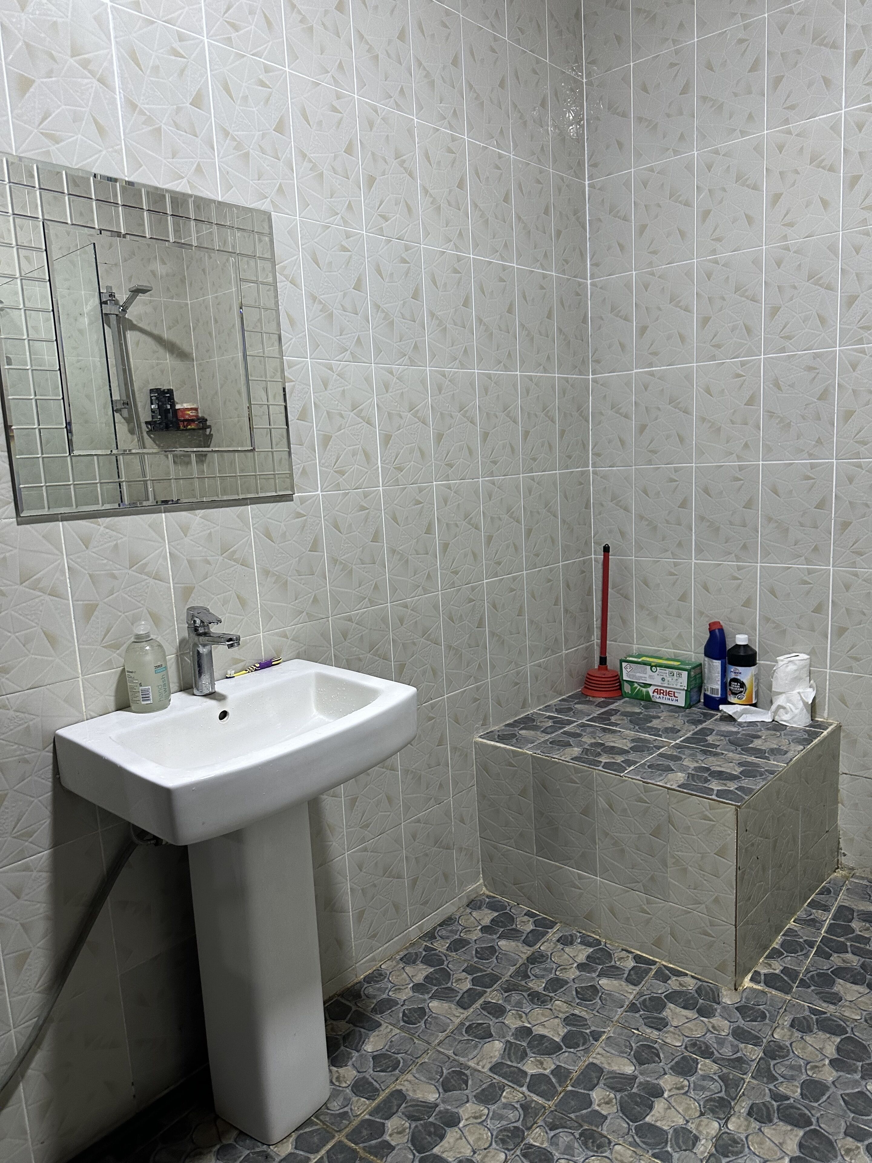A bathroom with a white sink, a mirror, tiled walls, a shower, and a small storage area with toiletries.