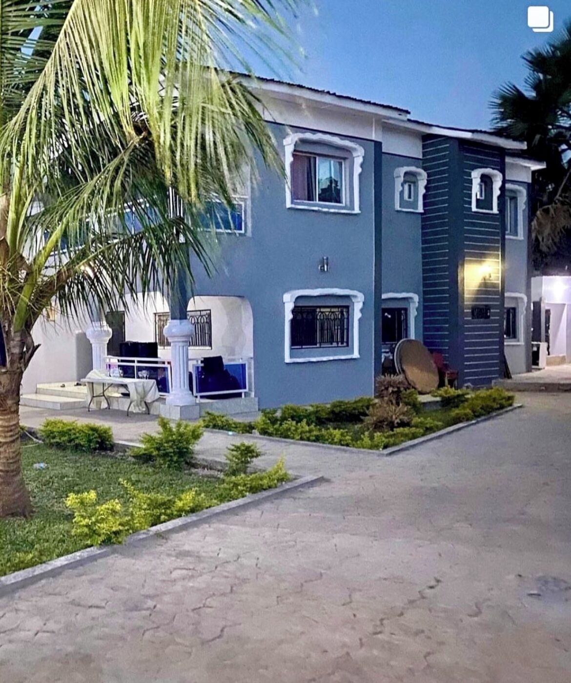 Un edificio de dos pisos con techo plano, un balcón y un pequeño jardín con una palmera.