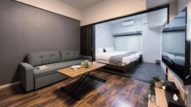 Una habitación moderna y funcional con un sofá gris, una mesa de centro de madera, una cama con cabecera gris y un televisor sobre un mueble.