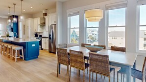 A modern kitchen with white cabinets, a blue island, stainless steel appliances, and a dining area with wicker chairs and a large window. - Blue Oasis - New + Modern w/Ocean Views, Pool + Hot Tub, 2 Min Walk to Beach, Elevator, Dogs OK (Carolina Beach)