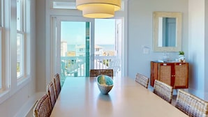 A dining area with a white table, wicker chairs, a hanging light fixture, and a view through glass doors to the outside. - Blue Oasis - New + Modern w/Ocean Views, Pool + Hot Tub, 2 Min Walk to Beach, Elevator, Dogs OK (Carolina Beach)