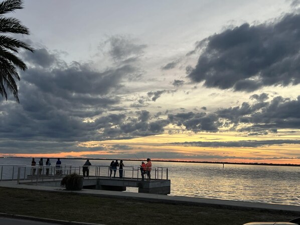 A waterfront promenade with a metal railing, people walking, and a sunset in the background.