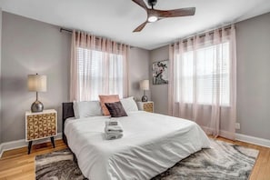 Quarto moderno com uma cama grande, duas mesas de cabeceira, um ventilador de teto e cortinas leves.