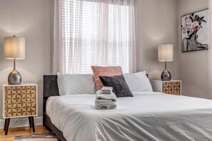 A neatly made bed with white bedding, two bedside lamps, a decorative wall piece, and a geometric-patterned nightstand.