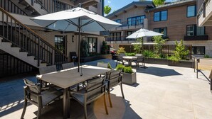 En udendørs patio med spisebord og stole, omgivet af krukker med planter og parasoller.