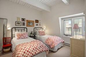 Une chambre avec deux lits, un fauteuil rouge, une petite table avec une lampe et une fenêtre avec des stores.