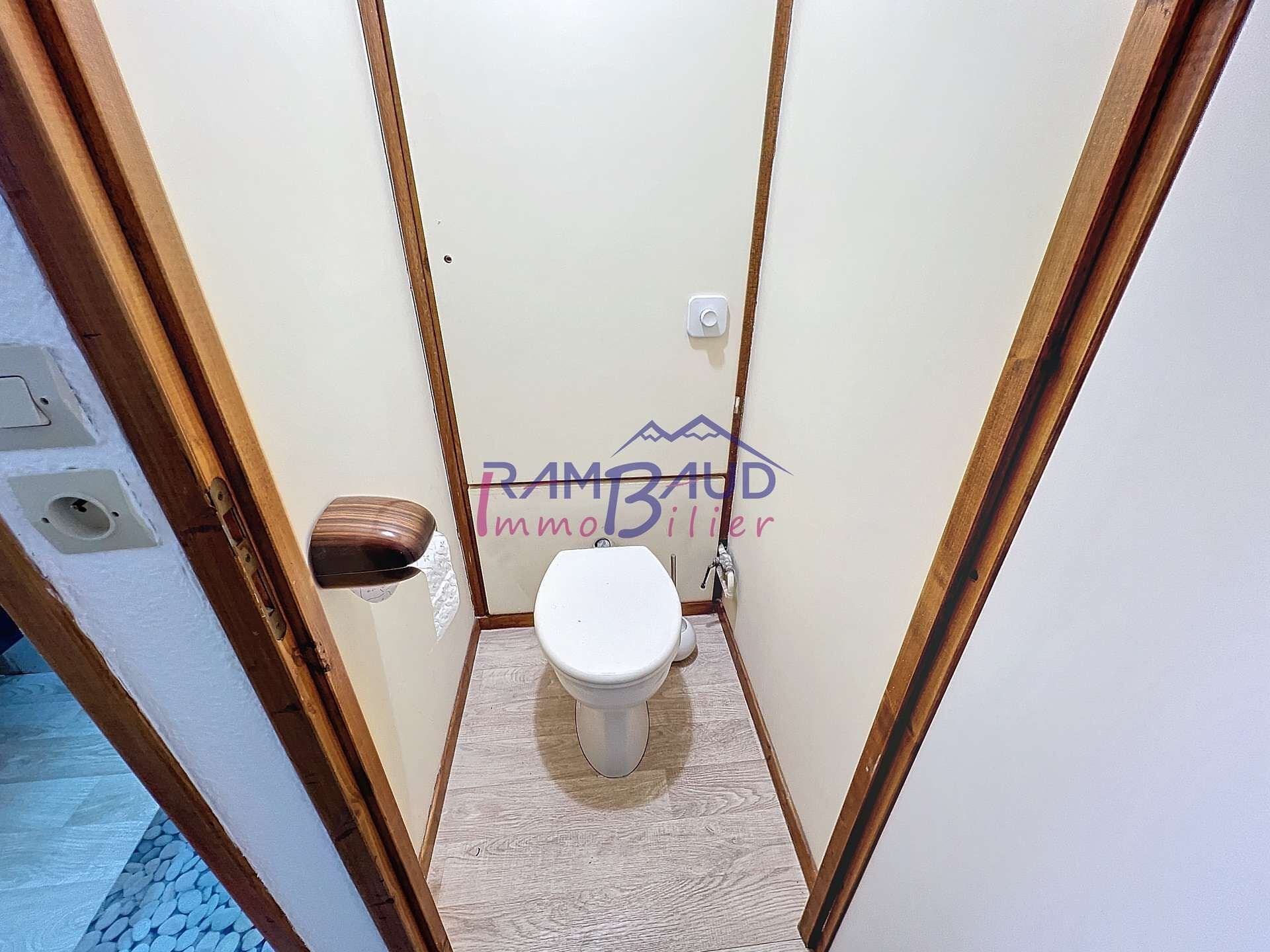 Une petite salle de bain étroite, avec des toilettes blanches, un cadre de porte en bois et une prise électrique visible.