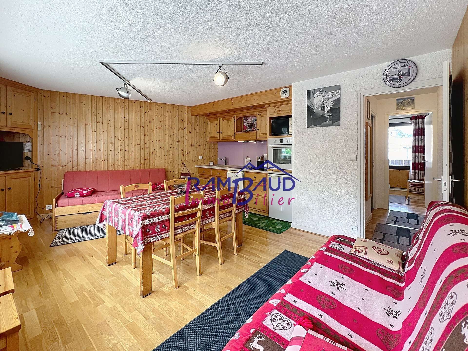 Una sala de estar acogedora con un sofá de madera, una mesa de comedor con sillas, una zona de cocina y un televisor pequeño.
