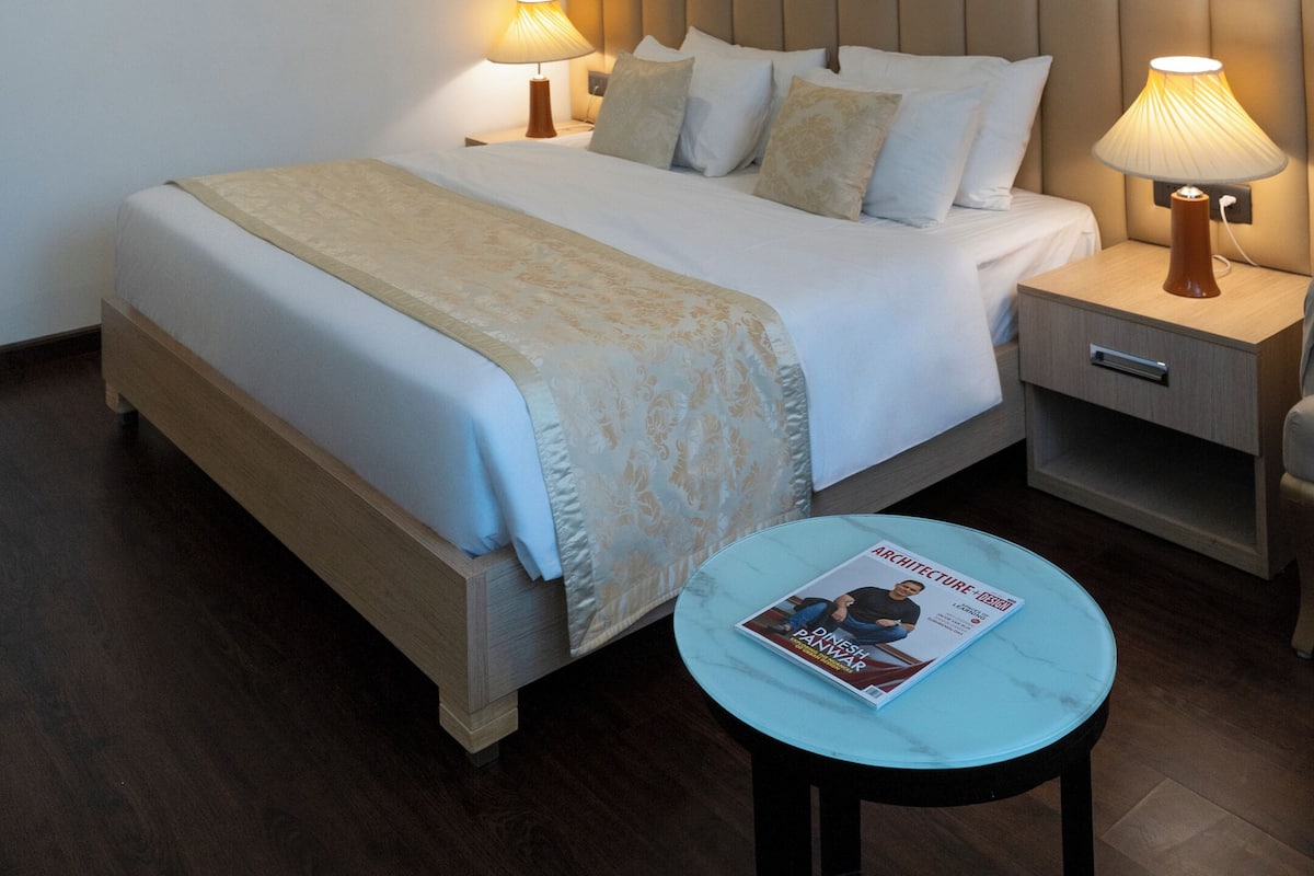 A hotel room with a large bed, two bedside tables with lamps, a small round table with a magazine, and two framed pictures on the wall.