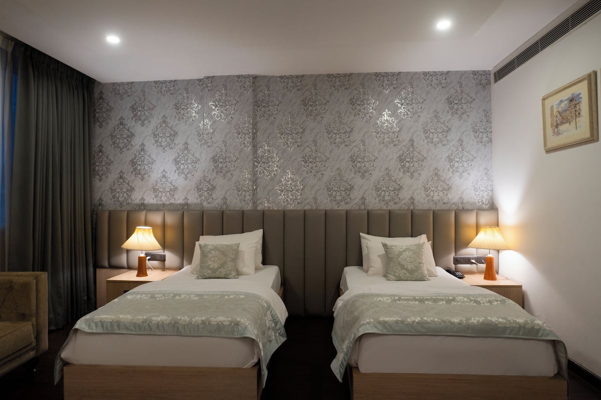 A hotel room with two beds, a headboard with a patterned wallpaper, bedside lamps, and a chair.