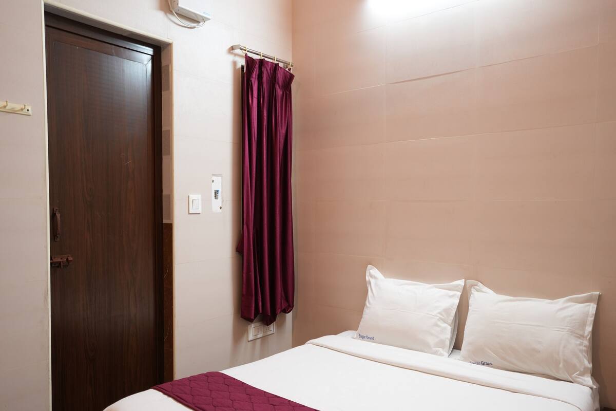 Quarto de hotel com uma cama, porta de madeira, luminária na parede e tomada.